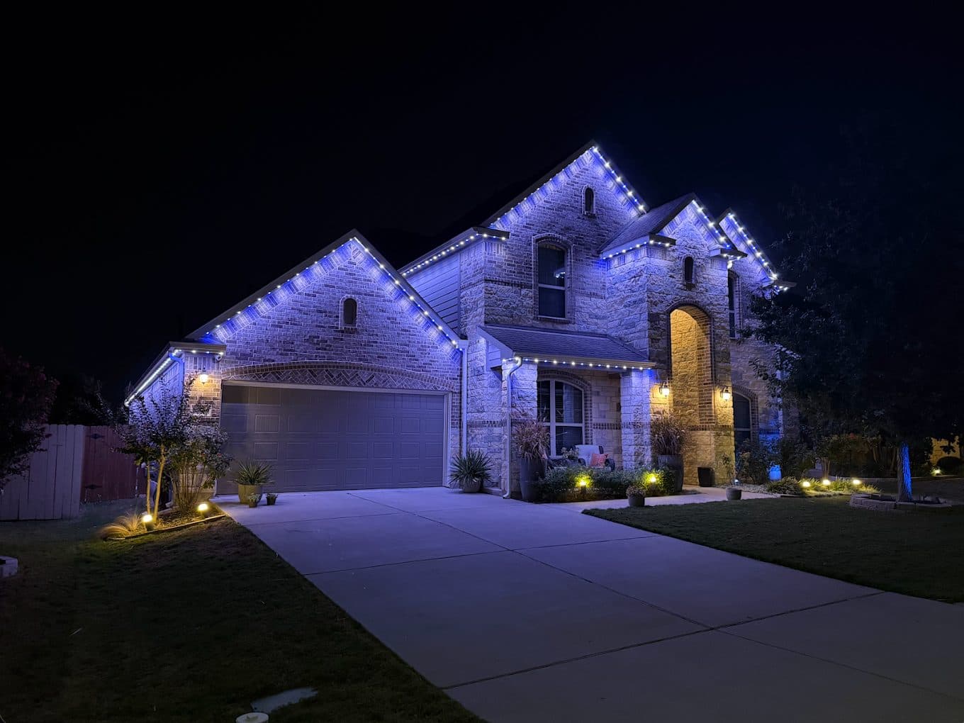 A stone multi-gable Elmhurst home with its permanent roofline lighting set to a blue accent scene, landscape fixtures lit below